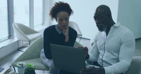 Pointing man, leaning woman collaborating on curved lounge chair, with laptop and coffee cup