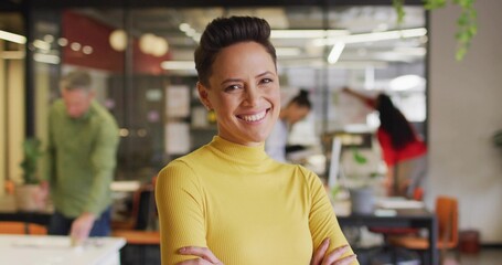 Smiling woman wearing yellow turtleneck crossing arms in open office with laptops and glass wall