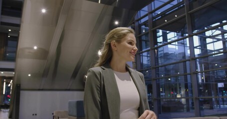 Standing woman wearing grey blazer in office lobby, escalator, glass wall, benches, recessed lights