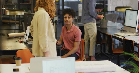 Hispanic man and female coworker reviewing blueprints in open-plan office, with laptop and monitors