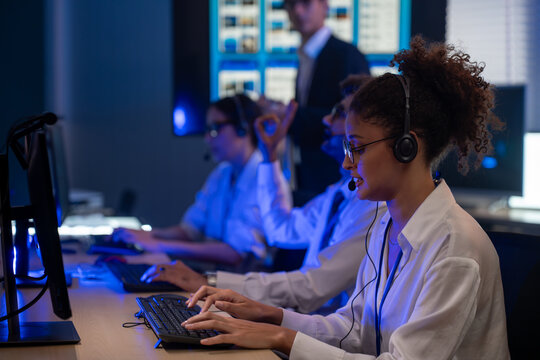 Diverse team of professionals customer service agent wearing headsets and monitoring computer working in dark control room. Female support and assistance call center at night in modern office.