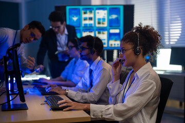 Diverse team of professionals customer service agent wearing headsets and monitoring computer working in dark control room. Female support and assistance call center at night in modern office.