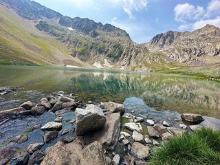 Pristine high-altitude mountain lake surrounded by rocky peaks and patches of snow. The water reflects the mountains and the cloudy blue sky. Large stones and green grass in the foreground.