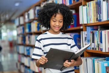 Smiling African American young student girl reading book while standing near bookshelf in school library. Learning, knowledge, and education concepts.