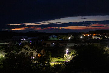 Fototapeta premium Scenic village at twilight with glowing streetlights and dramatic clouds above the horizon. Peaceful rural atmosphere with mountain silhouettes and colorful sunset sky in the background