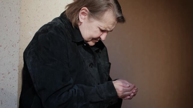 A miserable, poor elderly woman begs for alms, pressed against a wall