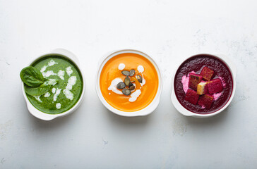 Three different bowls of healthy cream soups, broccoli peas green, orange pumpkin, and red beet, with fresh ingredients top view white background