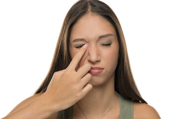 Fototapeta premium Studio shot of young woman with long brown hair wearing green tank top, scratching her eye with eyes closed, on white background.