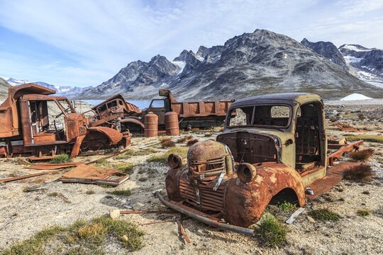 Rusty cars and oil drums in front of steep mountains, remains of a US airbase from the Second World War, Ikateq Fjord, East Greenland, Greenland