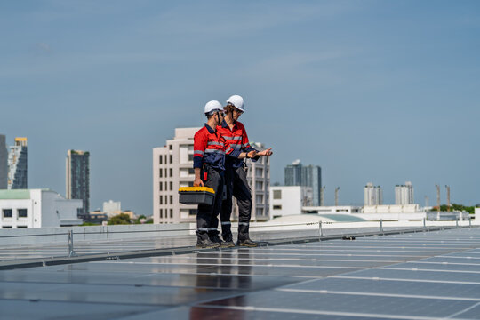 Solar engineer teamwork maintenance energy rooftop panel blue sky, two technician hard hat inspect array with tool box and tablet, urban background shows modern building and sustainable focus