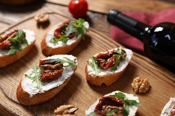 Delicious bruschettas with cream cheese, sun-dried tomatoes, arugula and bottle of wine on wooden table, closeup
