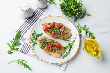 Delicious bruschettas with tuna, tomatoes and arugula served on white marble table, flat lay