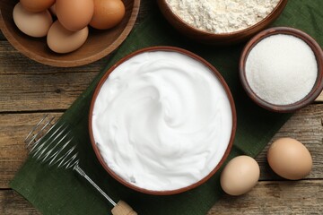 Whipped egg whites in bowl, whisk and ingredients on wooden table, flat lay