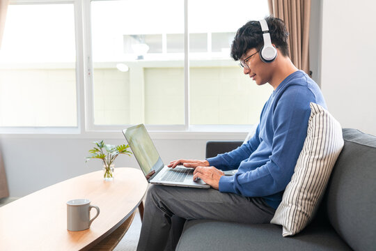 Asian man in casual clothes, typing business data on laptop and wearing headphone to listening music