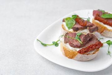 Bruschettas with roast beef, cream cheese, basil and sun-dried tomatoes on light grey table, closeup. Space for text