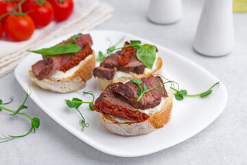 Bruschettas with roast beef, cream cheese, basil, sun-dried tomatoes and fresh vegetables on light grey table, closeup
