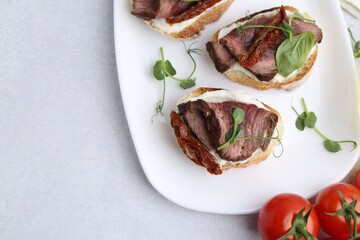 Bruschettas with roast beef, cream cheese, basil, sun-dried tomatoes and fresh vegetables on light grey table, flat lay. Space for text