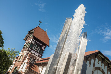 Moderner Springbrunnen aus poliertem Metall mit Wasserfontäne vor denkmalgeschützter alter Turnhalle mit Fachwerk und Walmdach in Metzingen. 