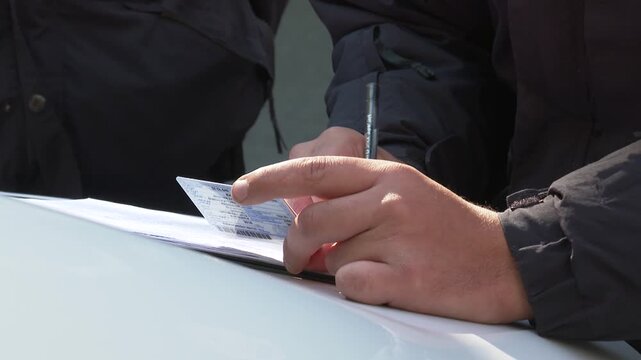 Close-up of police officer's hands writing an official report or traffic ticket, referencing an ID card on the hood of a car. Law enforcement procedure.