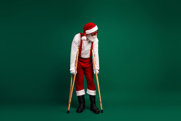Santa Claus dressed traditionally holds crutches while posing against an emerald green background, combining festive cheer with humor.