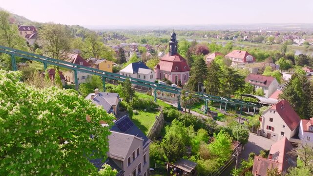 Drone flight over the Dresden Bergbahn Schwebebahn mountain railway during the day. The camera flies toward a train moving uphill, surrounded by greenery, trees, and a few houses.