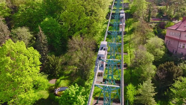 Drone flight over the Dresden Bergbahn Schwebebahn  mountain railway during the day. The camera moves to the left as two trains pass each other, surrounded by greenery, trees, and houses.