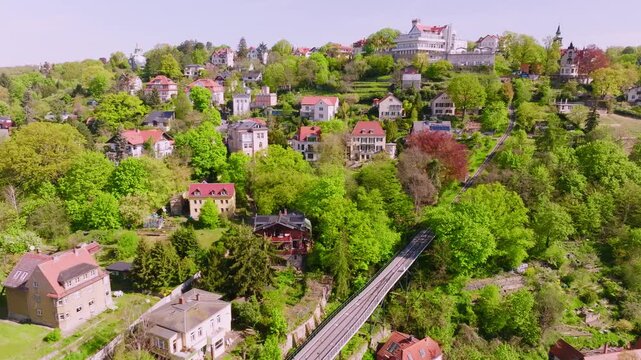 Drone flight over the Dresden Bergbahn Schwebebamountain railway during the day. The camera passes by the train, with the tracks clearly visible for a long stretch, surrounded by greenery and houses.
