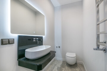 A simple and elegant bathroom design with a sink bowl on a stone countertop, mirrored lighting and a wall-mounted toilet.