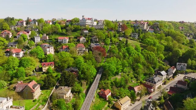 Drone flight over the Dresden mountain railways during the day. Two trains pass each other as the camera moves from right to left above houses, trees, and the wooded hillside.