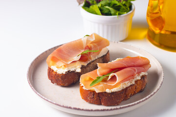 Two bruschettas with prosciutto, ricotta cheese and rucola on ciabatta bread. Olive and spices  decorated.