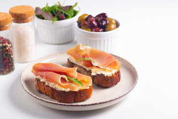 Two bruschettas with prosciutto, ricotta cheese and rucola on ciabatta bread. Olive and spices  decorated.