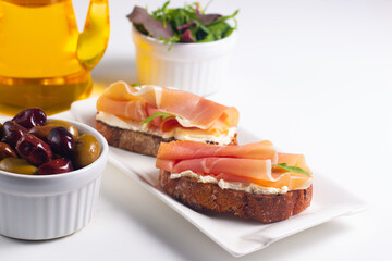 Two bruschettas with prosciutto, ricotta cheese and rucola on ciabatta bread. Olive and spices  decorated.