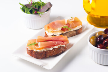 Two bruschettas with prosciutto, ricotta cheese and rucola on ciabatta bread. Olive and spices  decorated.
