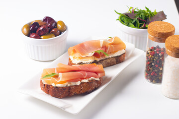 Two bruschettas with prosciutto, ricotta cheese and rucola on ciabatta bread. Olive and spices ...