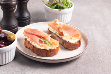 Two bruschettas with prosciutto, ricotta cheese and rucola on ciabatta bread. Olive and spices  decorated.