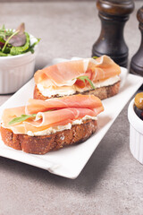 Two bruschettas with prosciutto, ricotta cheese and rucola on ciabatta bread. Olive and spices  decorated.