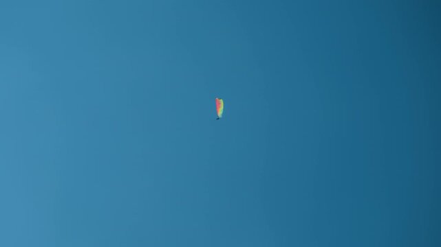 Upward shot of a man flying on a rainbow-colored paraglider against a clear blue sky, capturing aerial adventure and freedom.