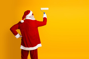Santa Claus painting a yellow wall with a roller while wearing his traditional red Christmas attire...