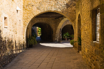 Fototapeta premium Stone archways and cobbled path in Monells, Catalonia, with potted plants and warm sunlight creating a peaceful medieval ambiance.