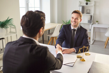 Smiling friendly lawyer in a suit shaking hands with a client at his office table with scales of...