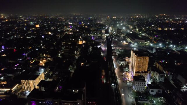 Aerial drone moving forward shot showing train crossing over elevated metro bridge and busy street filled with lights and lit cars underneath with cityscape of indian city of jaipur in distance