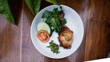 A plate of Indonesian food featuring fried chicken, cucumber, tomato, and a side of sambal.