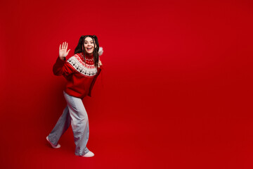 Young woman in a festive red Christmas sweater waves cheerfully against a vivid red background perfect for holiday campaigns and seasonal fashion visuals