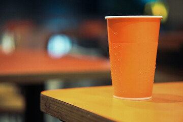Orange paper cup with condensation on a table in a casual dining setting. Cold beverage container in a restaurant atmosphere with blurred background and warm lighting.