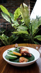 A delicious dish of grilled chicken with fresh cucumber and sambal served on a banana leaf in a garden setting.