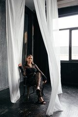 Confident woman in sheer black lace stands poised between white curtains, embodying strength, elegance, and modern gothic fashion
