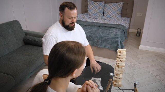 Young couple is playing a game of jenga in their living room, carefully removing blocks from the tower and placing them on top, trying not to make it fall