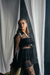 Confident woman in sheer black lace stands poised between white curtains, embodying strength, elegance, and modern gothic fashion