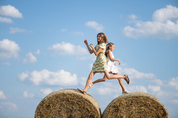 Two young girls, one in a floral dress and the other in a white dress, joyfully jumping between hay bales in a sunny field, capturing the essence of childhood playfulness and freedom © shymar27