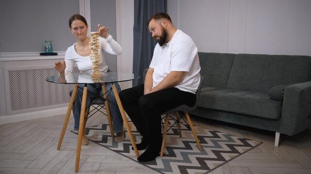 Young couple is playing a game of jenga in their living room, carefully removing blocks from the tower and placing them on top, trying not to make it fall